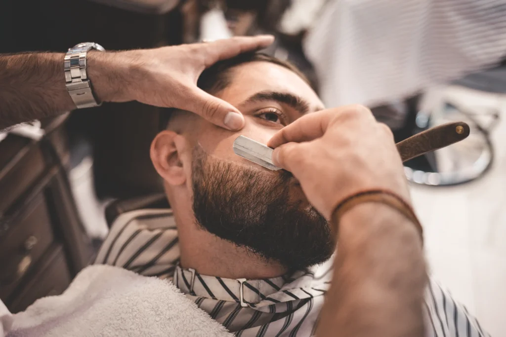 Barber shaping a client's beard with a straight razor.